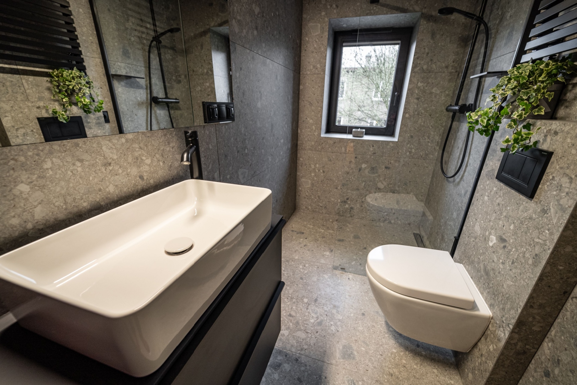 Moderne bad med mørke overflater. Veggene og gulvet er i mørke steinfliser, hvitt WC, sort dusjapparat, sort benk hvor servanten står, stort speil og en plante på en hylle over WC